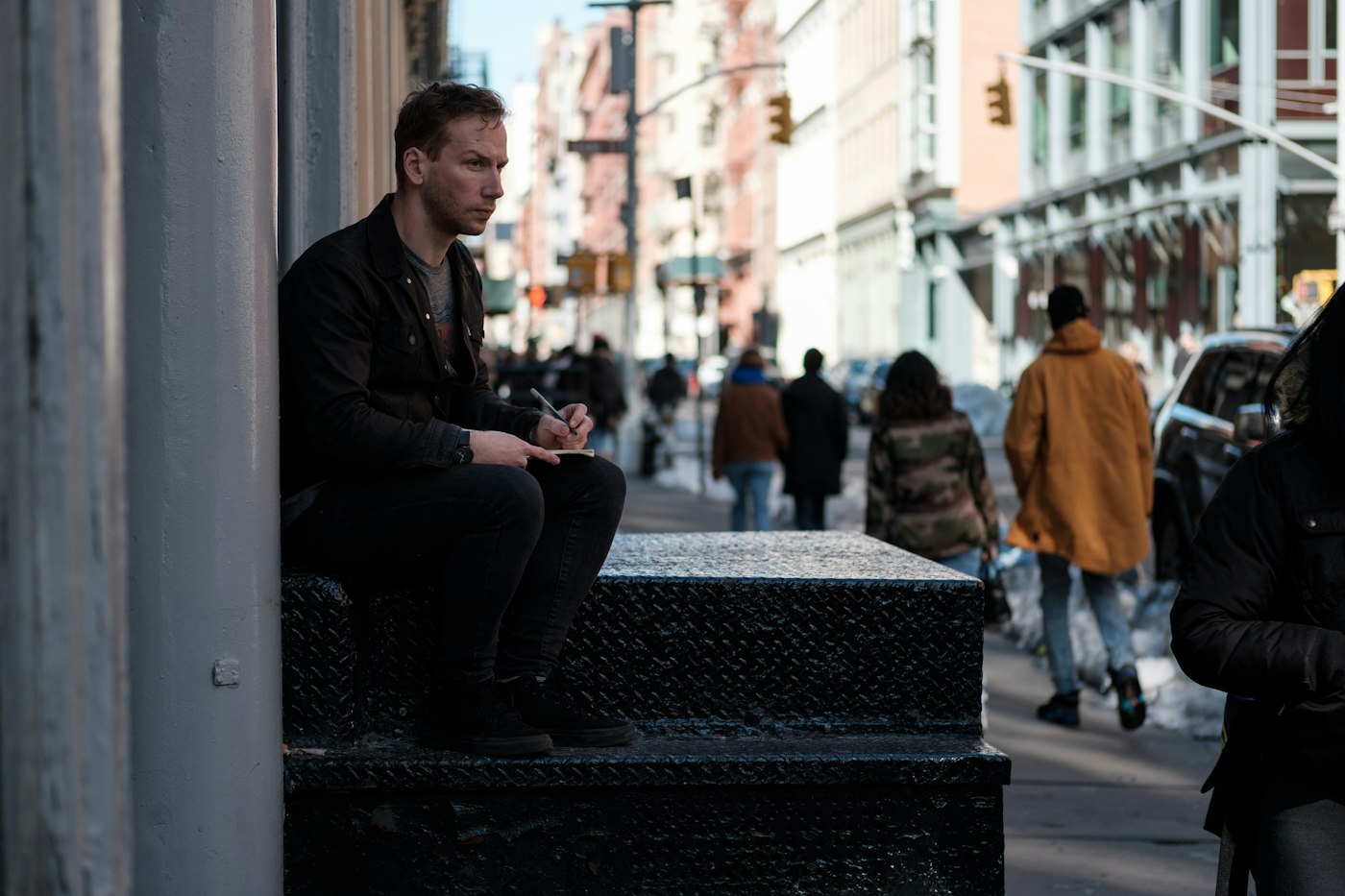 Man sitting on a stone ledge with a sketchpad on a busy city sidewalk