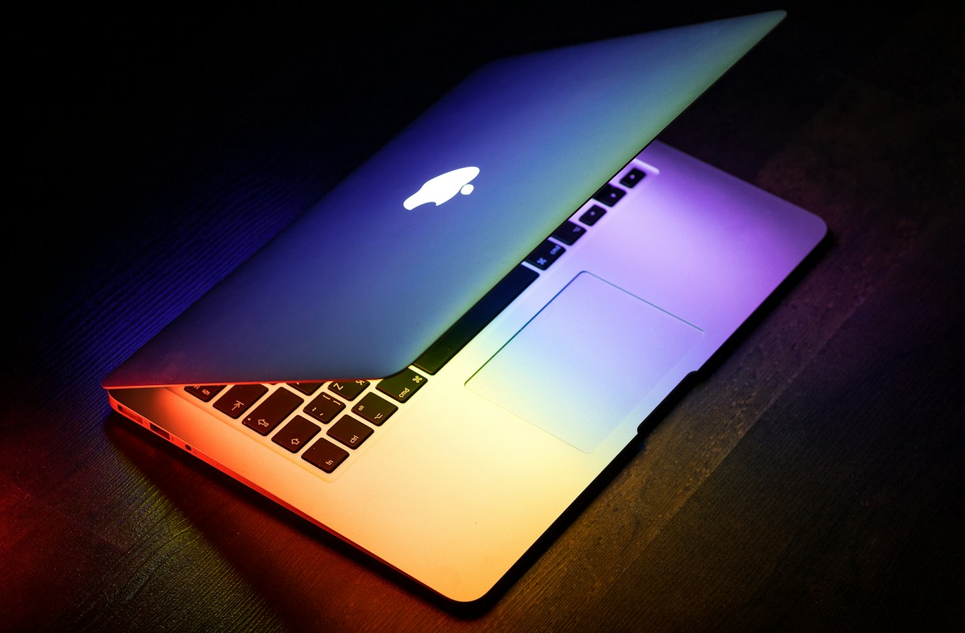 Half-open MacBook glowing with rainbow light on a dark wooden table